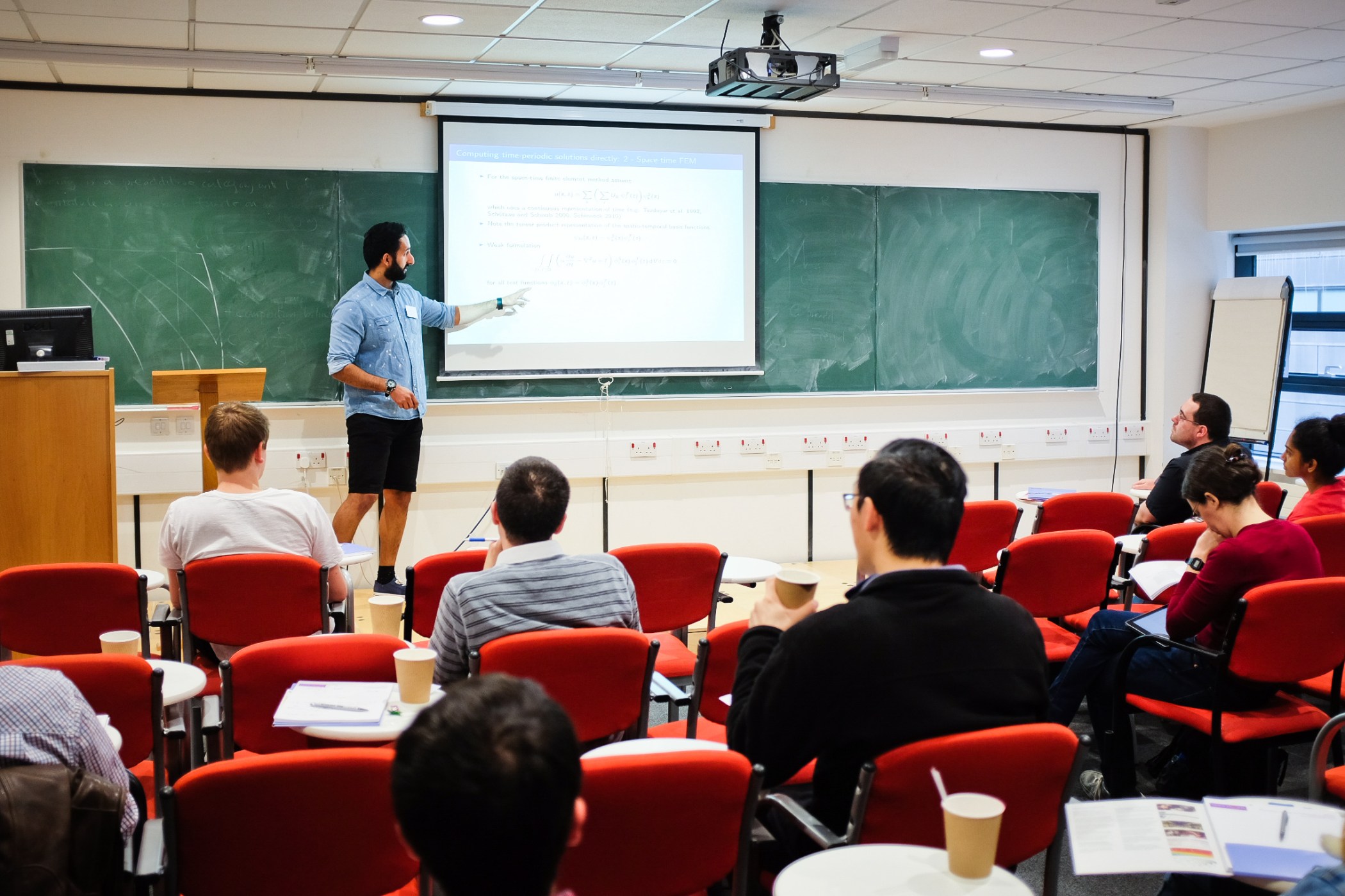 A mathematics talk in the Frank Adams room, with blackboards, a projection screen and red audience seating.
