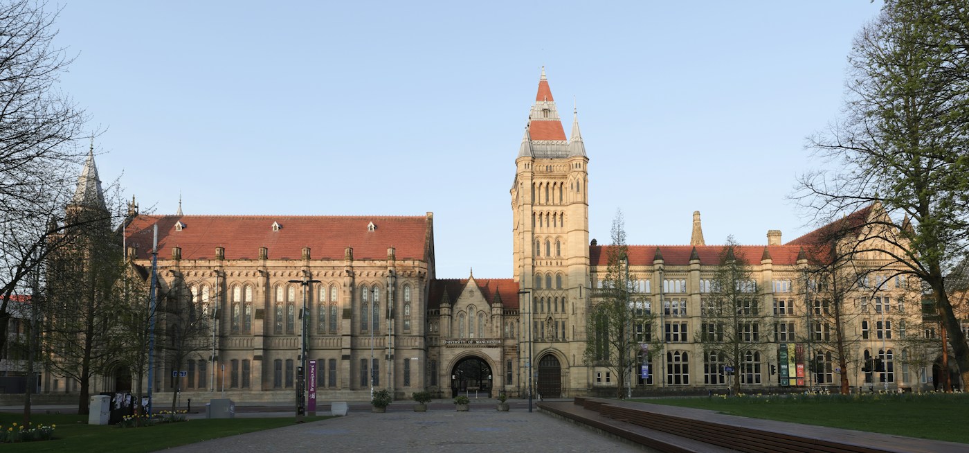 Historic University of Manchester buildings, with a tall stone tower and red roofs, viewed across the courtyard.